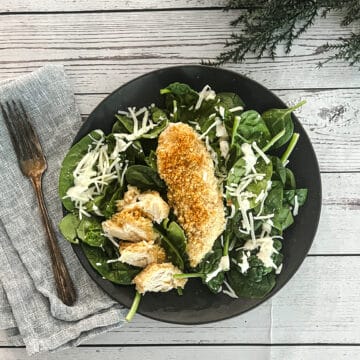 A plate of salad with chicken and spinach on a wooden table. The chicken contains parmesan cheese, caesar croutons, and caesar dressing.