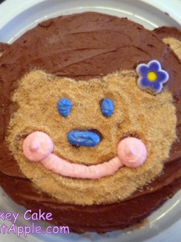 A round cake with a monkey-shaped face made of brown frosting and graham cracker crumbs. The cake has two purrple frosting eyes, a pink nose, and a mouth made of pink frosting.