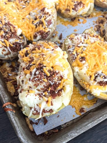 A close-up photo of twice baked potatoes on a baking sheet. The potatoes are golden brown and crispy on top, and filled with a creamy mashed potato mixture. The mashed potatoes are topped with shredded cheese and bacon