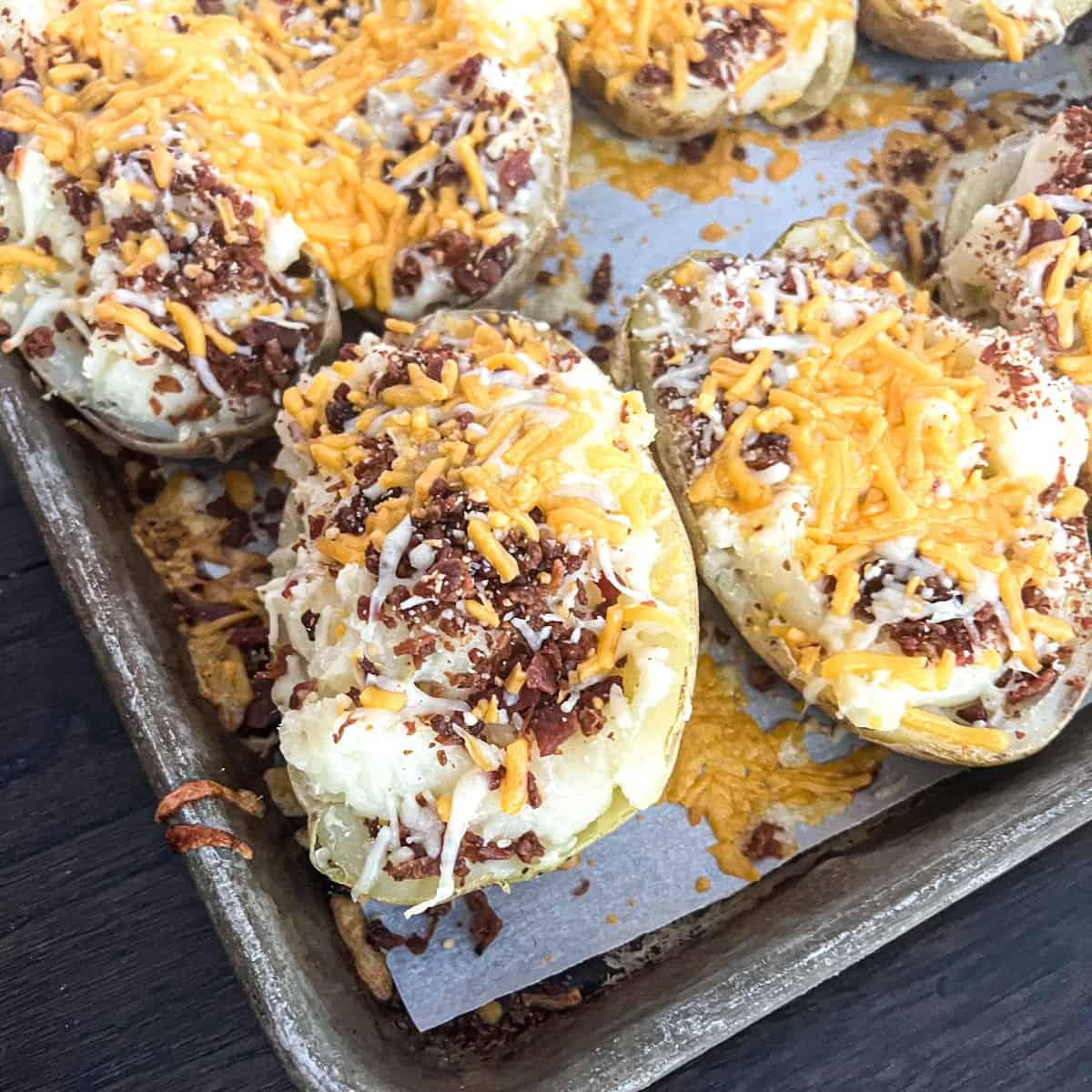 A close-up photo of  twice baked potatoes on a baking sheet. The potatoes are golden brown and crispy on top, and filled with a creamy mashed potato mixture. The mashed potatoes are topped with shredded cheese and bacon