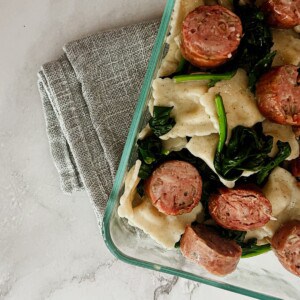a photo of butternut squash ravioli, apple chicken sausage and spinach in a clear dish placed on a gray napkin.
