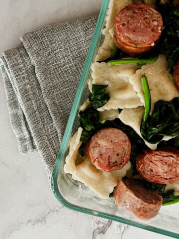 a photo of butternut squash ravioli, apple chicken sausage and spinach in a clear dish placed on a gray napkin.
