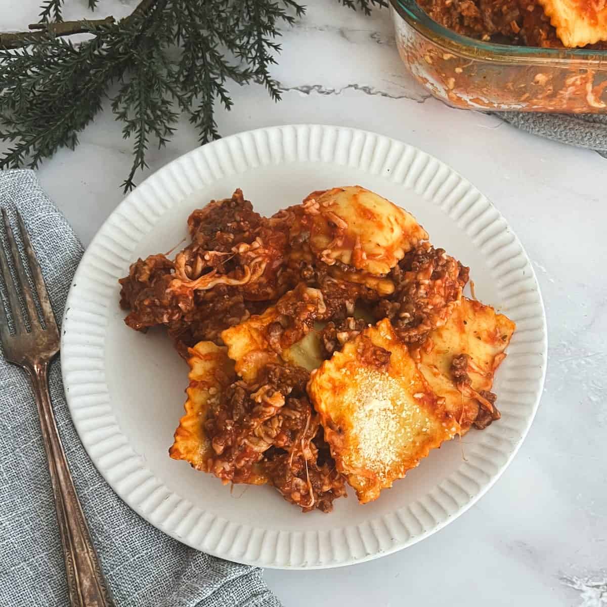 A  plate of ravioli bake with meat sauce and melted cheese on a table. The ravioli bake is on a white plate next to a casserole dish. The casserole dish is filled with ravioli, meat sauce, and melted cheese.