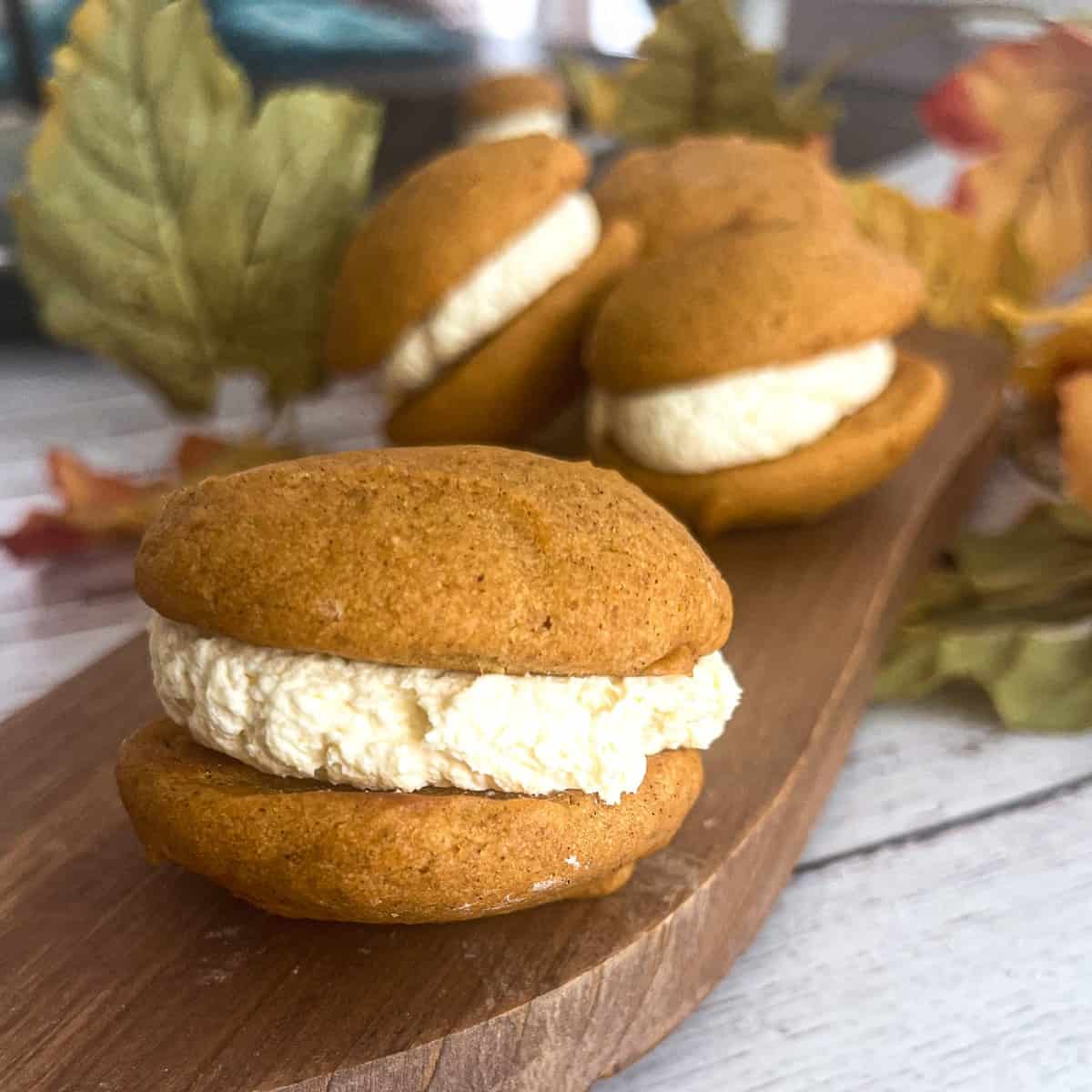  close-up photograph of two Pumpkin Whoopie Pies stacked on top of each other. The pies consist of two soft, cake-like cookies with a rich, creamy cream cheese frosting filling in between. The cookies are a deep, golden-brown color, and the frosting is generously spread between them.