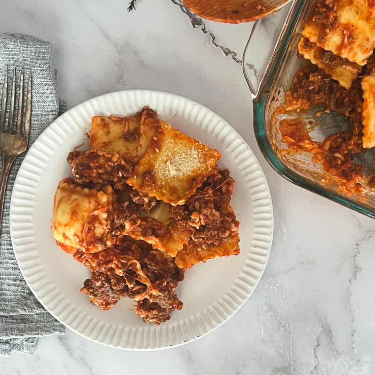 A plate of ravioli bake with meat sauce on a table. The ravioli casserole is on a white plate next to a casserole dish. The casserole dish is filled with ravioli, meat sauce, and melted cheese. The ravioli bake is topped with chopped fresh parsley.