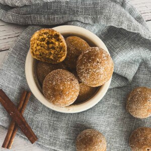 A bowl of pumpkin spice donut holes on a white table. The donut holes are golden brown and dusted with cinnamon sugar. A cinnamon stick is placed next to the bowl.