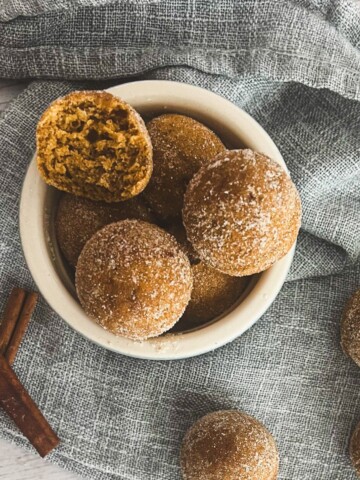A bowl of pumpkin spice donut holes on a white table. The donut holes are golden brown and dusted with cinnamon sugar. A cinnamon stick is placed next to the bowl.