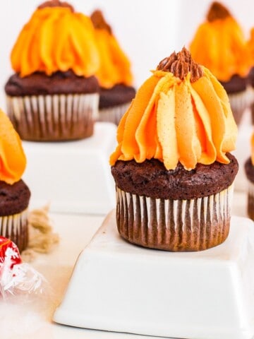 A close-up photo of a chocolate pumpkin cupcake on a white plate. The cupcake is topped with a generous swirl of buttercream frosting. The frosting is smooth and creamy, and the cupcake is moist and fluffy.