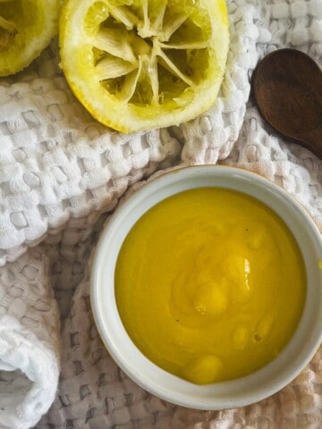 A close-up photo of a jar of Meyer lemon curd. The curd is a bright yellow color and has a smooth and silky texture. The jar is sitting on a white table, and there is a spoon next to it.