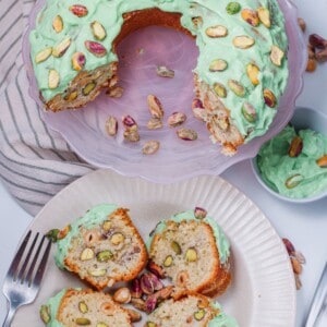 A close-up photo of a pistachio cake decorated with a thick and creamy pistachio cake frosting. The frosting is a light green color, speckled with finely ground pistachios, and has a smooth, piped swirl design on top of the cake. The cake itself is visible underneath, showing a moist, golden brown crumb with flecks of pistachios throughout.