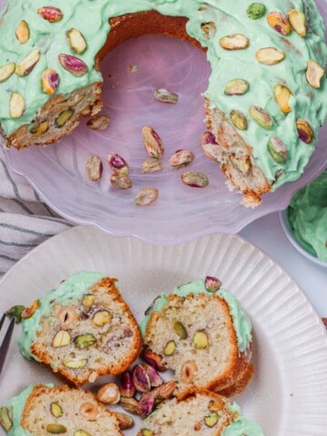 A close-up photo of a pistachio cake decorated with a thick and creamy pistachio cake frosting. The frosting is a light green color, speckled with finely ground pistachios, and has a smooth, piped swirl design on top of the cake. The cake itself is visible underneath, showing a moist, golden brown crumb with flecks of pistachios throughout.