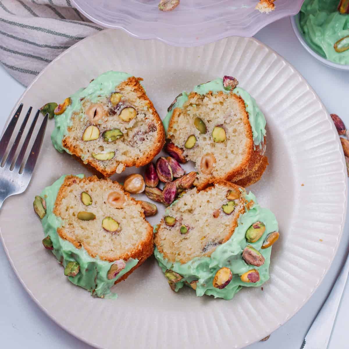 A close-up photo of a slice of pistachio pudding cake on a white plate. The cake is light and fluffy, with a moist, green interior due to the pistachio pudding mix. There is a generous swirl of white frosting on top, decorated with a sprinkle of chopped pistachios. The plate is garnished with a single whole pistachio nut