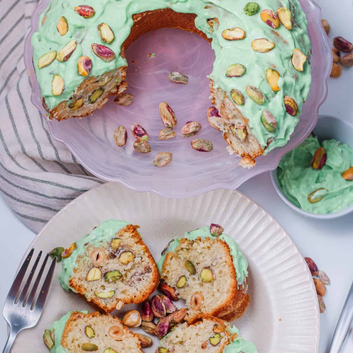 A close-up photo of a moist pistachio cake on a white plate. The cake is light and fluffy, with a light green swirl visible throughout the crumb. It has a golden brown, slightly cracked top sprinkled with chopped pistachios. A thick glaze coats the top of the cake, dripping slightly down the sides. A single whole pistachio nut sits on the edge of the plate for garnish