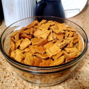 A glass bowl filled with gluten-free golden graham crackers on a counter. The crackers are square-shaped and have a light golden brown color. Some of the crackers are tilted on their sides, so you can see their thickness. There are also crumbs scattered on the counter around the bowl.