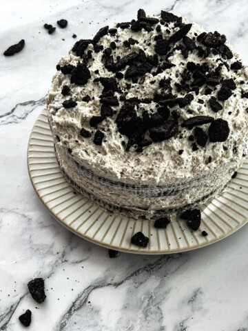 A close-up photo of a rich, chocolate-colored frosting swirled on top of a chocolate cake. The Oreo frosting recipe is generously studded with visible chunks of crushed Oreo cookies, adding a textural contrast. The cake is placed on a white plate, with a small fork resting beside it.
