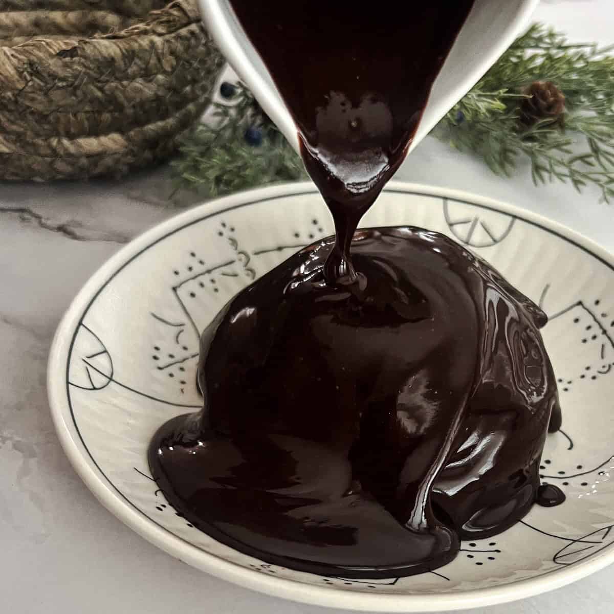 A small saucepan filled with melted chocolate sauce. The sauce is a dark, glossy brown. A whisk sits in the pan. The sauce is a 3 ingredient hot fudge sauce. 