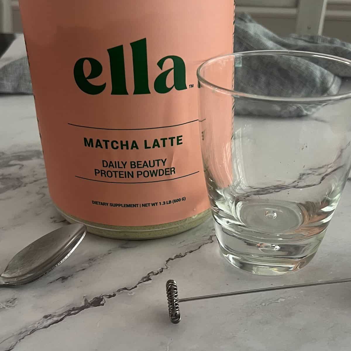 A glass of water sits next to a jar of matcha latte daily beauty protein powder. The jar has a black label with white and gold text. The text says "Naked Nutrition," "Ella" followed by "MATCHA LATTE," "DAILY BEAUTY," "PROTEIN POWDER," "DIETARY SUPPLEMENT