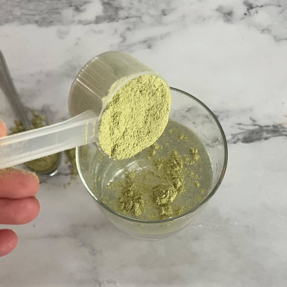 A close-up of a green powder being poured from a scoop into a glass of water. The powder is a bright green and appears to be finely ground.