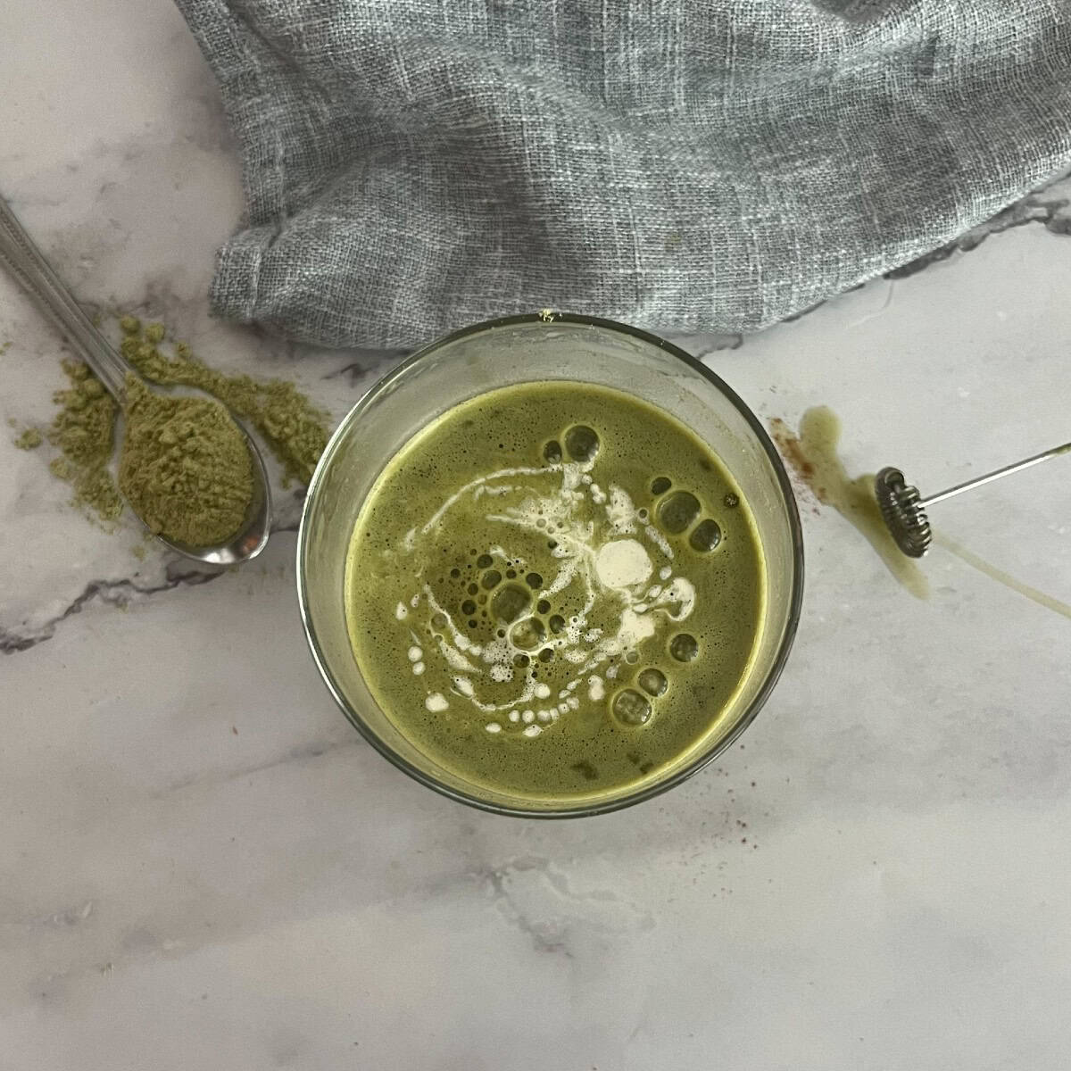 A close-up of a glass filled with a green matcha latte. The latte has a frothy layer on top and is garnished with a sprinkle of matcha powder on a marble countertop. Matcha powder is a finely ground green tea powder that is used in traditional Japanese tea ceremonies and other recipes.