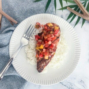 A white plate with a bed of white rice. On top of the rice is a boneless, skinless chicken breast that is cooked and cut into slices. The chicken is topped with a bright yellow pineapple mango salsa.
