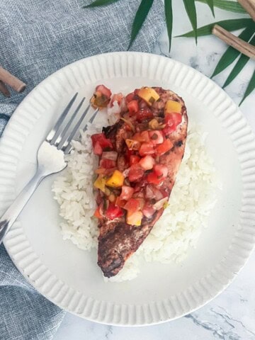 A white plate with a bed of white rice. On top of the rice is a boneless, skinless chicken breast that is cooked and cut into slices. The chicken is topped with a bright yellow pineapple mango salsa.