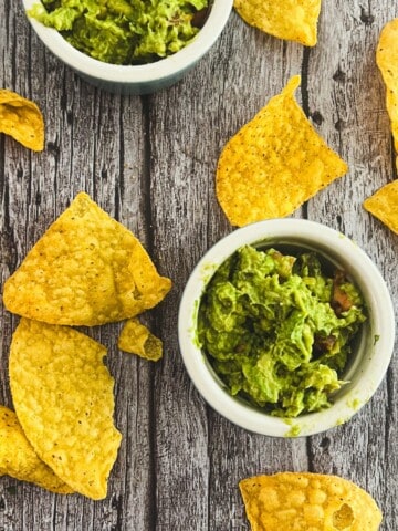 Two bowls of 4-ingredient guacamole recipe with a chunky texture. Tortilla chips are scattered around the bowls.