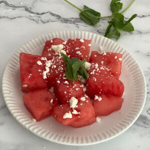 A white plate with sliced watermelon and goat cheese crumbles and mint leaves on top.