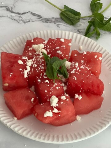 A white plate with sliced watermelon and goat cheese crumbles and mint leaves on top.