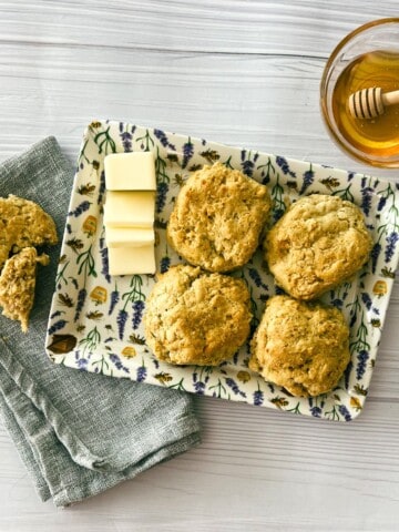 A plate of golden brown air fryer biscuits on a brown wooden table. The biscuits are round and flaky with a slight golden brown crust. There is a small dish of honey and a pat of butter on the side of the plate. In the background, there is a black air fryer on a black counter top.