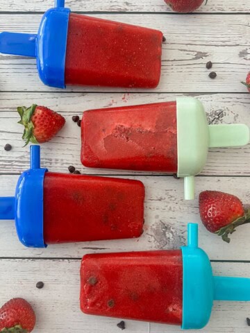 A close-up photo of four red popsicles with visible strawberry chunks. The popsicles are resting on a wooden table and text overlay says how to make strawberry popsicles