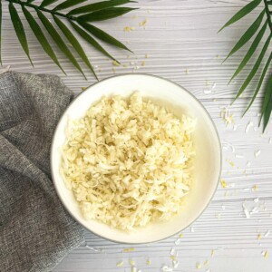 A white bowl filled with fluffy white rice, with a few pieces of lemon zest and shredded coconut sprinkled on top. The bowl is sitting on a white wooden table, with a gray napkin and a palm leaf in the background.