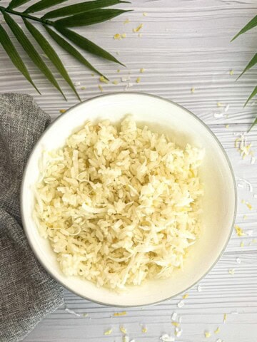 A white bowl filled with fluffy white rice, with a few pieces of lemon zest and shredded coconut sprinkled on top. The bowl is sitting on a white wooden table, with a gray napkin and a palm leaf in the background.