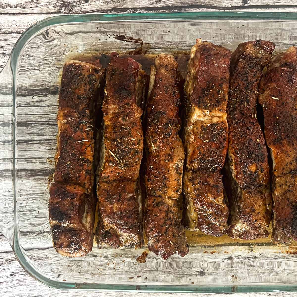 A glass baking dish filled with beef ribs in the oven. The ribs are a deep red color and appear to be cooked. There is a brown liquid with some fat pooling in the bottom of the dish. In the background, there is a blurred image of a wooden table