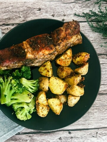 A black oval plate filled with crispy crispy garlic parmesan crusted potatoes. the potatoes are golden brown potato wedges. The potatoes have a rough, textured surface and are dusted with Parmesan cheese. In the background, there is a piece of steamed broccoli and a serving of ribs baked in the oven.