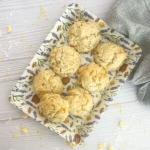 The image shows six lemon coconut cookies with a light and airy texture, arranged on a rectangular tray with a floral design. The cookies have a pale yellow hue and are generously sprinkled with white chocolate chips and shredded coconut. The tray is placed on a white wooden surface, with additional coconut flakes scattered around it.