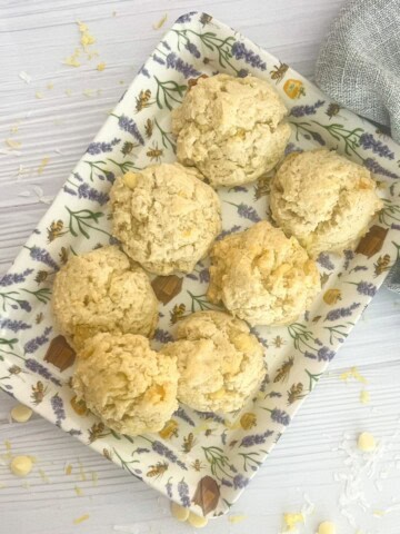 The image shows six lemon coconut cookies with a light and airy texture, arranged on a rectangular tray with a floral design. The cookies have a pale yellow hue and are generously sprinkled with white chocolate chips and shredded coconut. The tray is placed on a white wooden surface, with additional coconut flakes scattered around it.