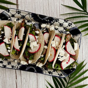 A plate of pork street tacos. Tacos: Soft tortillas filled with shredded pork, lettuce, and radishes. Toppings: The tacos are topped with crumbled feta cheese and additional radishes. Presentation: The tacos are arranged on a colorful plate with palm leaves and a wooden background, creating a visually appealing scene.