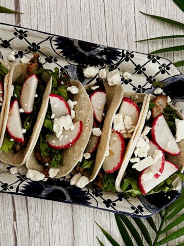 A plate of pork street tacos. Tacos: Soft tortillas filled with shredded pork, lettuce, and radishes. Toppings: The tacos are topped with crumbled feta cheese and additional radishes. Presentation: The tacos are arranged on a colorful plate with palm leaves and a wooden background, creating a visually appealing scene.