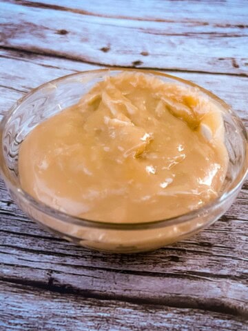 A small glass bowl filled with a thick 2 ingredient carmael sauce that is a caramel-colored sauce. The sauce has a smooth texture with a few ripples on the surface. It's sitting on a wooden table.