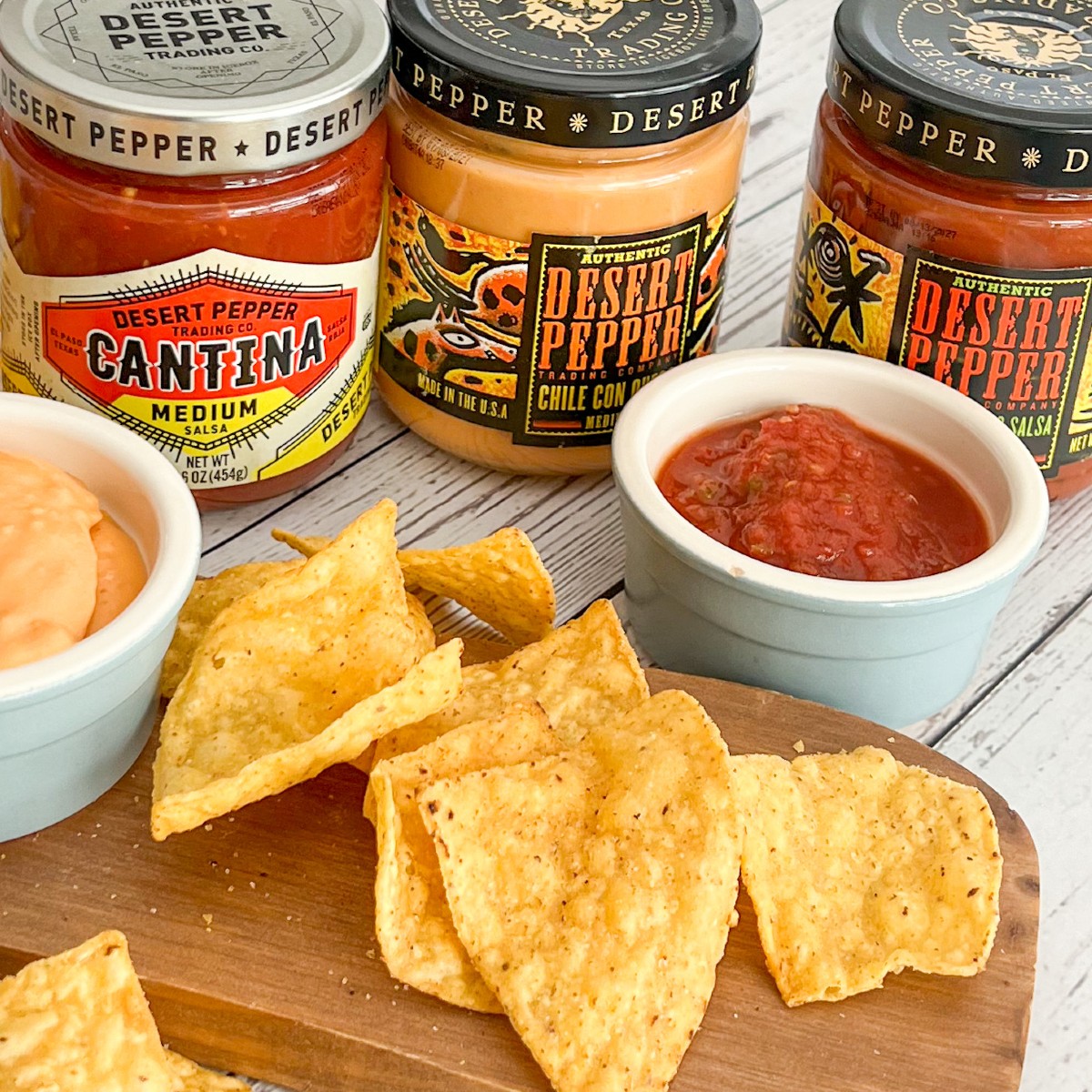 Three jars of Desert Pepper Trading Co. salsa, labeled "Cantina Medium," "Chile Con Carne," and "Salsa." The jars are arranged around a small bowl of queso dip and a scattering of tortilla chips on a wooden cutting board.