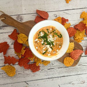 A white bowl filled with a creamy, orange queso dip with ground beef topped with crumbled feta cheese and chopped chives. The dip is surrounded by a variety of colorful tortilla chips.