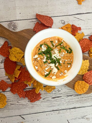 A white bowl filled with a creamy, orange queso dip with ground beef topped with crumbled feta cheese and chopped chives. The dip is surrounded by a variety of colorful tortilla chips.