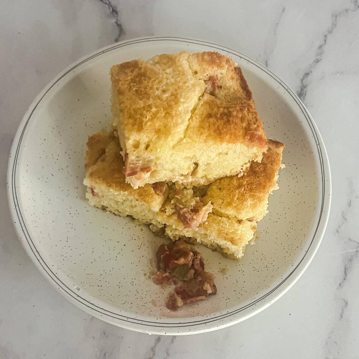 A plate of three squares of rhubarb dump cake mix. The cake is golden brown and has a crumbly texture. A small amount of rhubarb is visible on the plate.