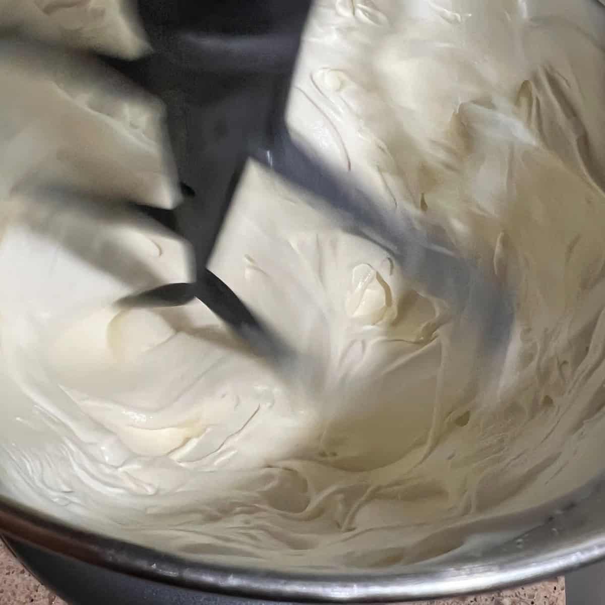 A close-up image of a mixer beating frosting in a silver bowl. The frosting is thick and creamy, with the mixer's blades visible as they mix it.