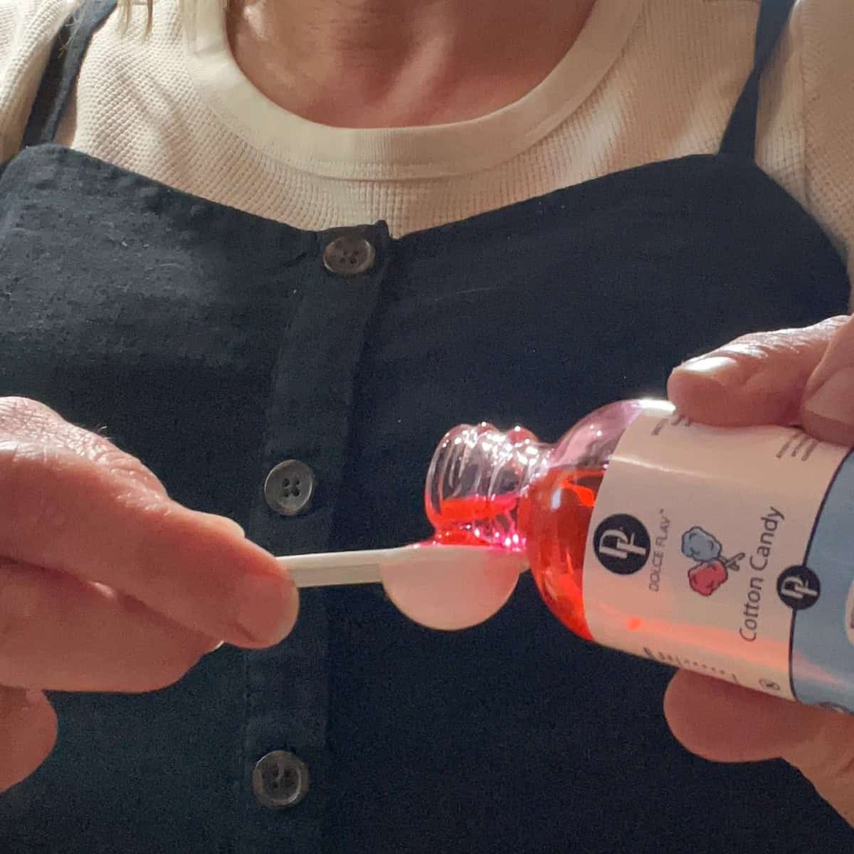 A person's hands holding a small, clear bottle of cotton candy flavoring. They are using a spoon to measure out a portion of the pink liquid. The bottle has a white label with "Dolce Flav" and "Cotton Candy" written on it.