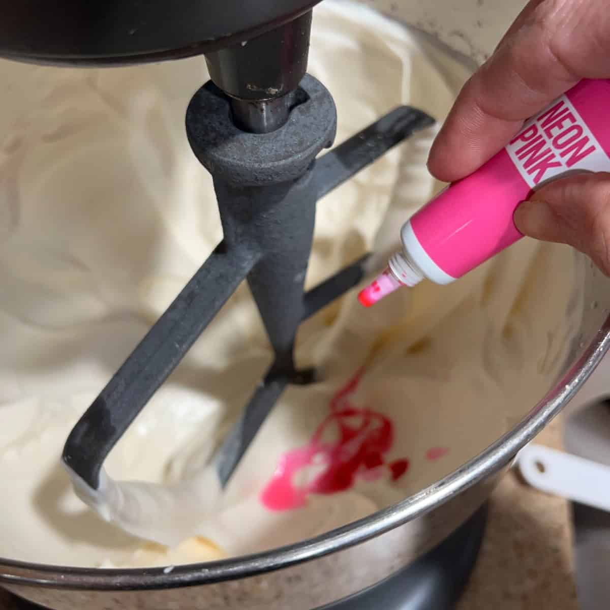 A close-up image of a hand squeezing a tube of neon pink food coloring into a bowl of white frosting. The frosting is being mixed by a mixer, and the pink color is starting to spread through the white.