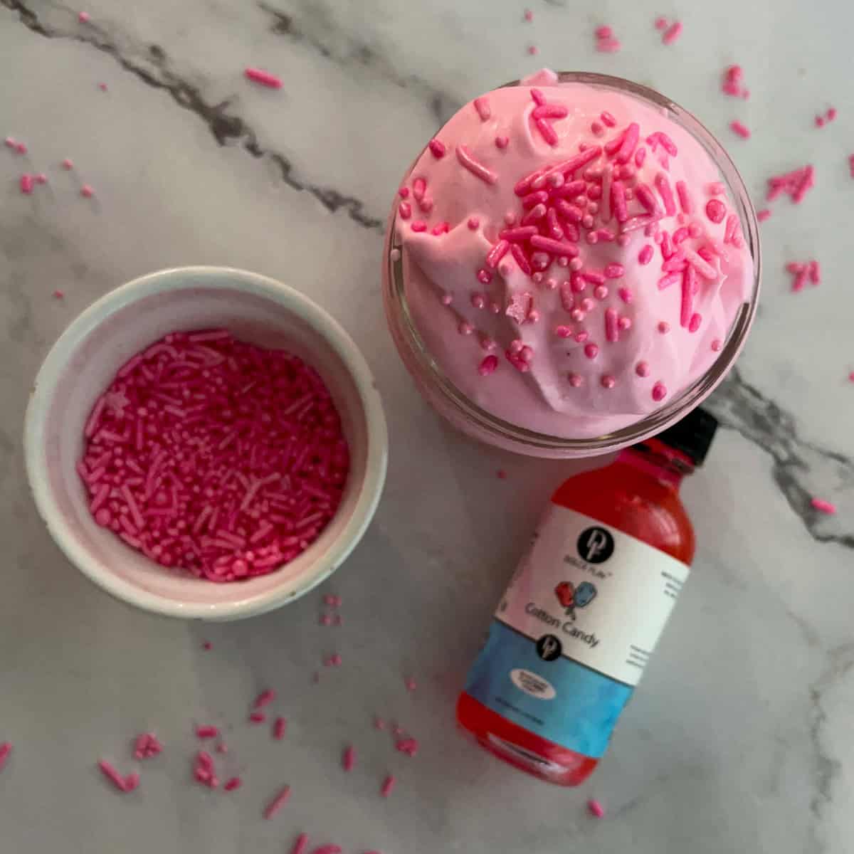 A mason jar filled with pink whipped cream topped with pink sprinkles. Next to the jar is a small bowl of pink sprinkles and a bottle of cotton candy flavoring.