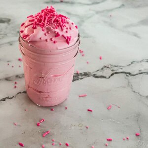 A mason jar filled with a recipe for cotton candy pink whipped cream topped with pink sprinkles. The cream is light and fluffy, with a hint of pink coloring. A few sprinkles are scattered on the surface and around the jar.