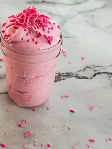 A mason jar filled with a recipe for cotton candy pink whipped cream topped with pink sprinkles. The cream is light and fluffy, with a hint of pink coloring. A few sprinkles are scattered on the surface and around the jar.