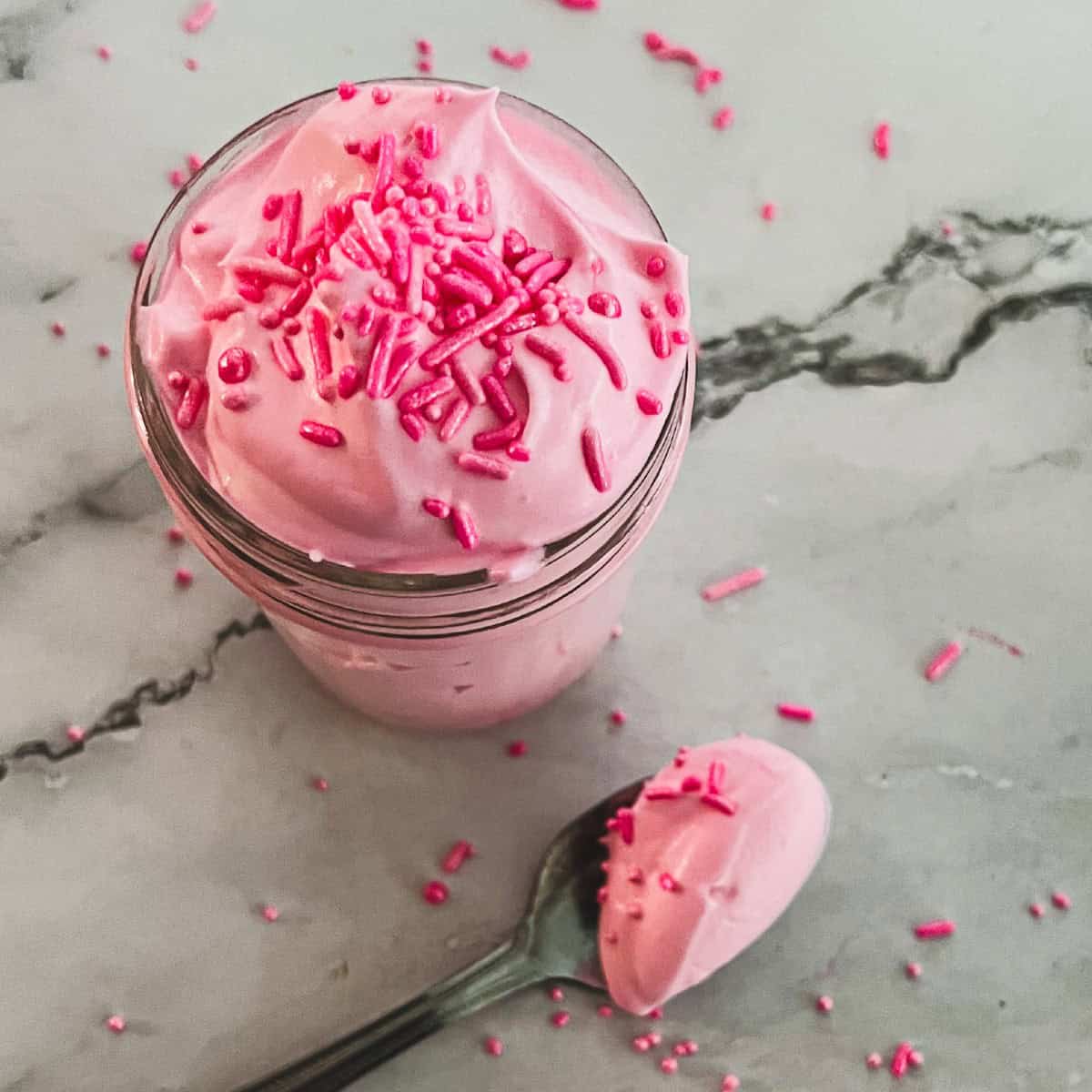 A mason jar filled with pink whipped cream topped with pink sprinkles. A spoon is dipped into the cream, with a dollop on the spoon. This is a recipe for cotton candy flavoring. 